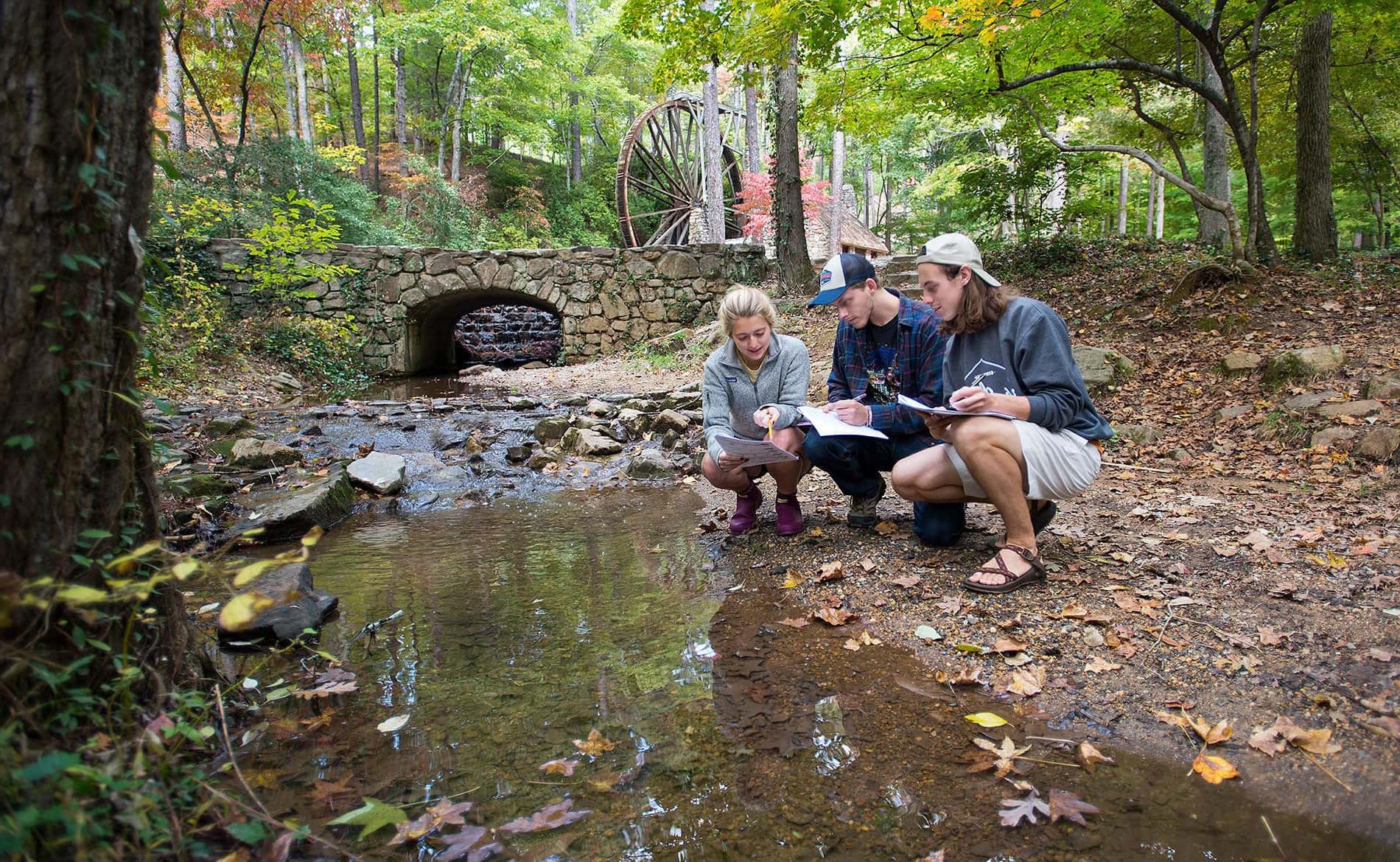 three students kneeling by a creek examinig papers